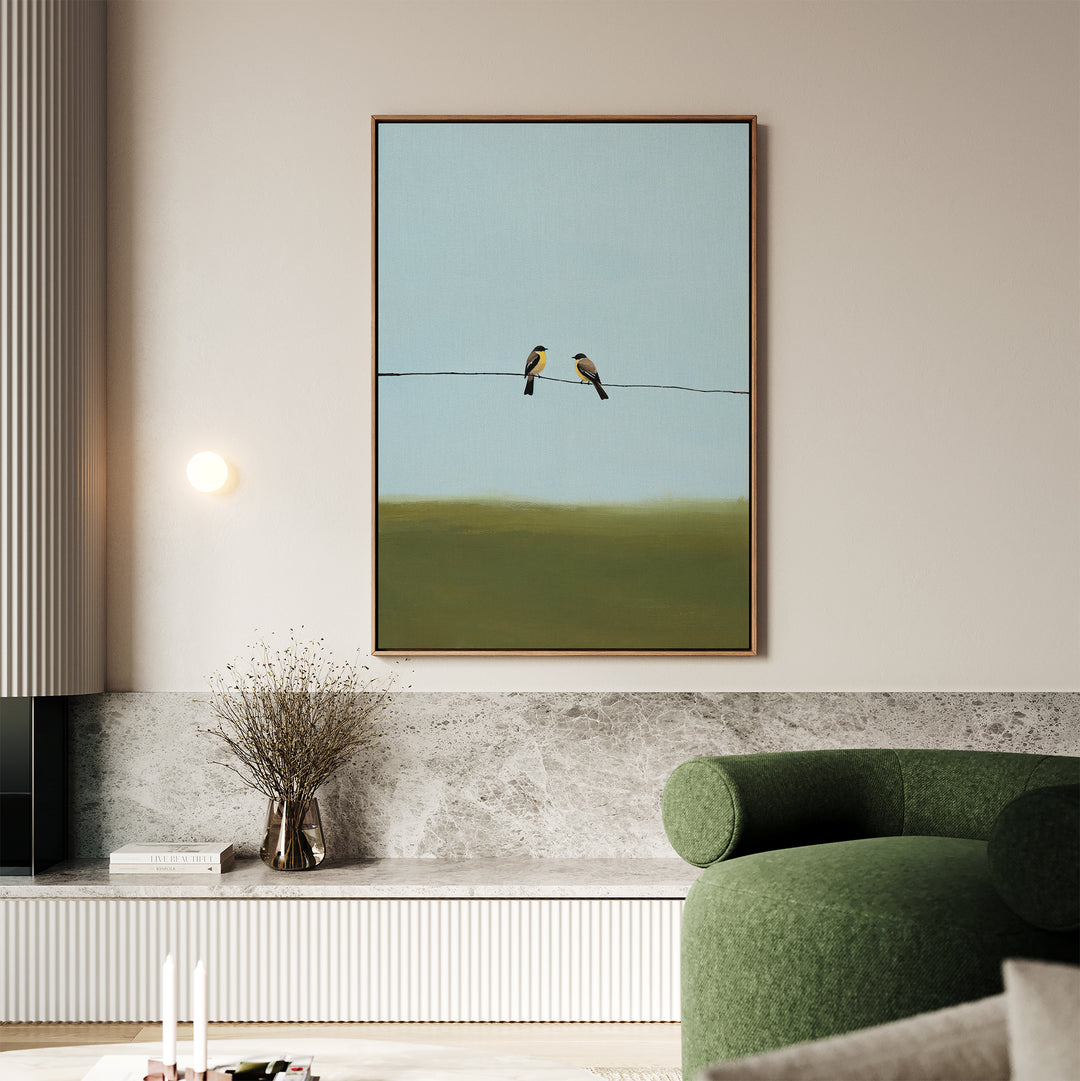 A modern living room displays the Meadow Line Canvas Art, a minimalist print of two birds on a wire, elegantly hung above a marble shelf with dried branches and complemented by a green curved sofa in the foreground.