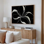 A modern living room featuring a light wood sideboard, books, a lamp, neutral decor, and the Midline Motion Canvas Art—a striking minimalist black-and-white floral painting—hanging on the wall above.