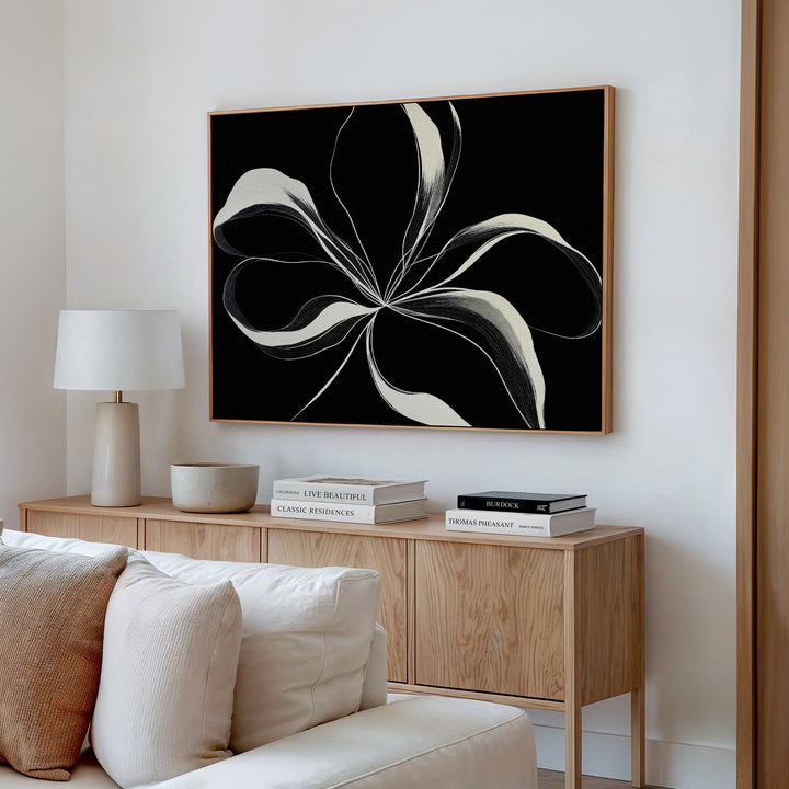 A modern living room featuring a light wood sideboard, books, a lamp, neutral decor, and the Midline Motion Canvas Art—a striking minimalist black-and-white floral painting—hanging on the wall above.