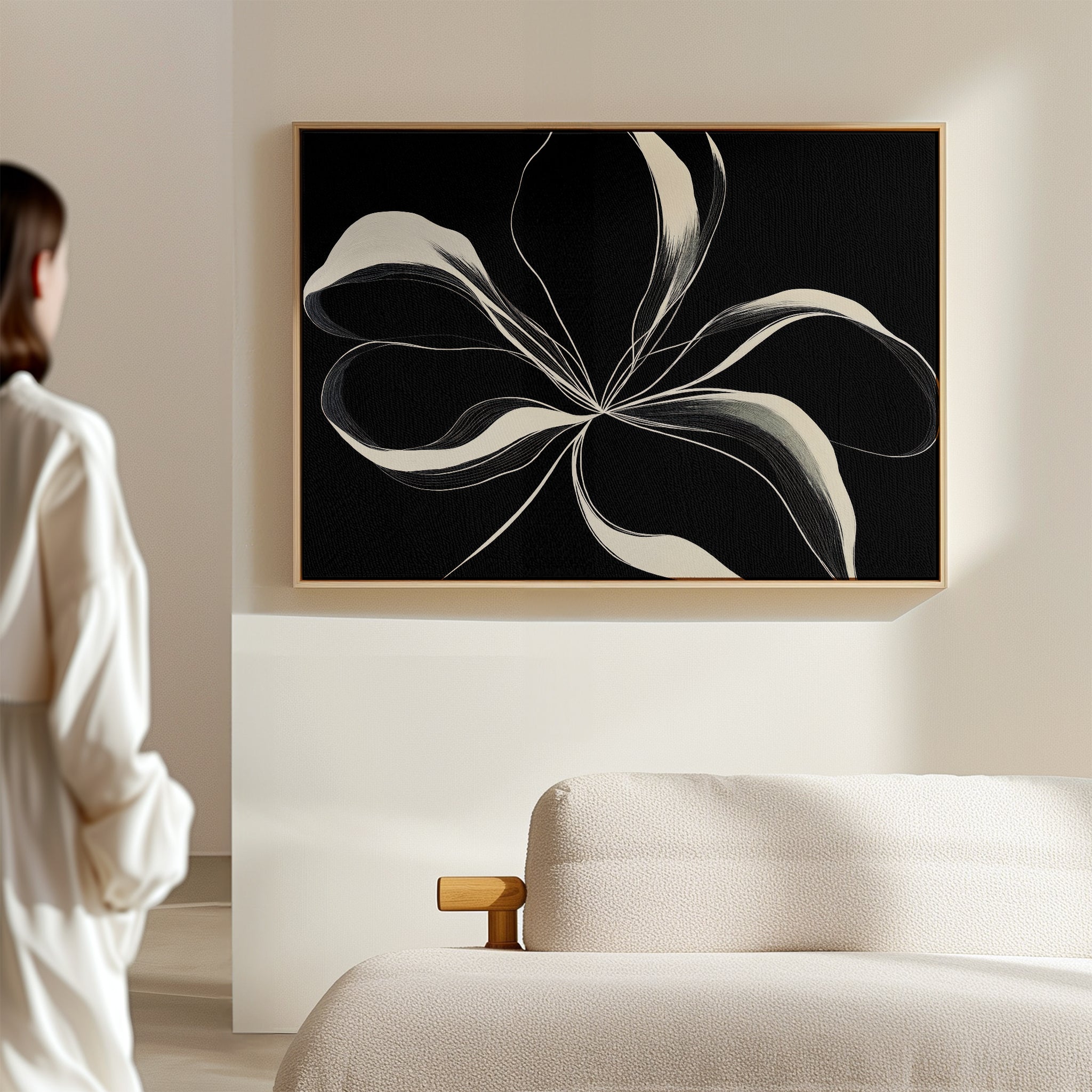 In a minimal, light-filled room, a person admires the Midline Motion Canvas Art—a large abstract black-and-white floral piece displayed above a cream-colored sofa—adding the perfect touch of modern living room decor.