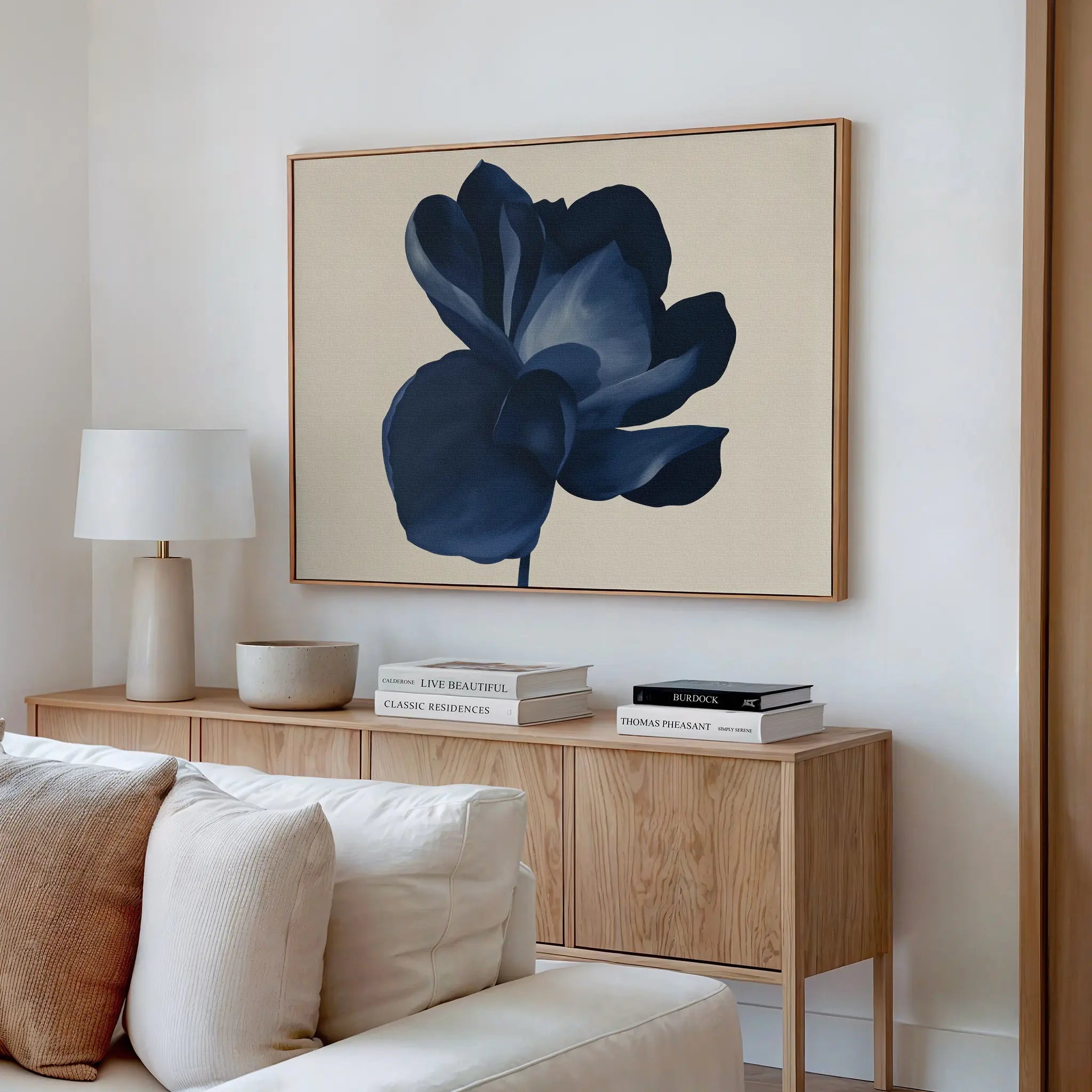 A modern living room features a light wood sideboard with books, a round lamp, and ceramics. Above it, the Midnight Bloom Canvas Art – Horizontal brings sophisticated, minimalist flair. A cream sofa sits in the foreground.