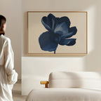 A woman in a white robe stands in a minimal, sunlit room, admiring the Midnight Bloom Canvas Art – Horizontal, whose large blue flower design adds modern sophistication above the beige upholstered bed.