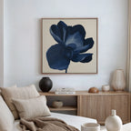 A modern living room features a beige sofa, neutral cushions, wooden sideboard, decorative vases, stacked books, and a large Midnight Bloom Canvas Art – Square showcasing a dark blue flower on the wall.