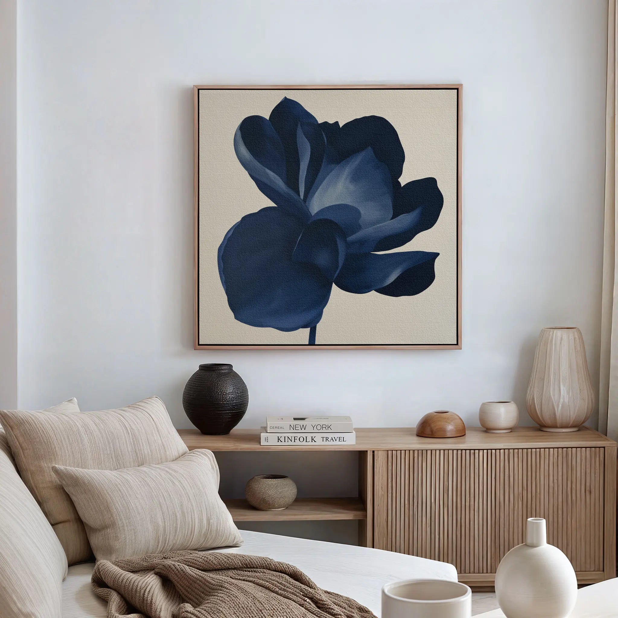 A modern living room features a beige sofa, neutral cushions, wooden sideboard, decorative vases, stacked books, and a large Midnight Bloom Canvas Art – Square showcasing a dark blue flower on the wall.