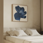 A beige bedroom features a tufted headboard, white bedding, and the Midnight Bloom Canvas Art – Square with a large blue flower above the bed. A bare branch in a vase decorates the right side.