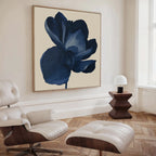 A modern living room featuring a white leather lounge chair with ottoman, a wooden side table with a small white lamp, and the Midnight Bloom Canvas Art – Square, displaying a dark blue flower on the beige wall.