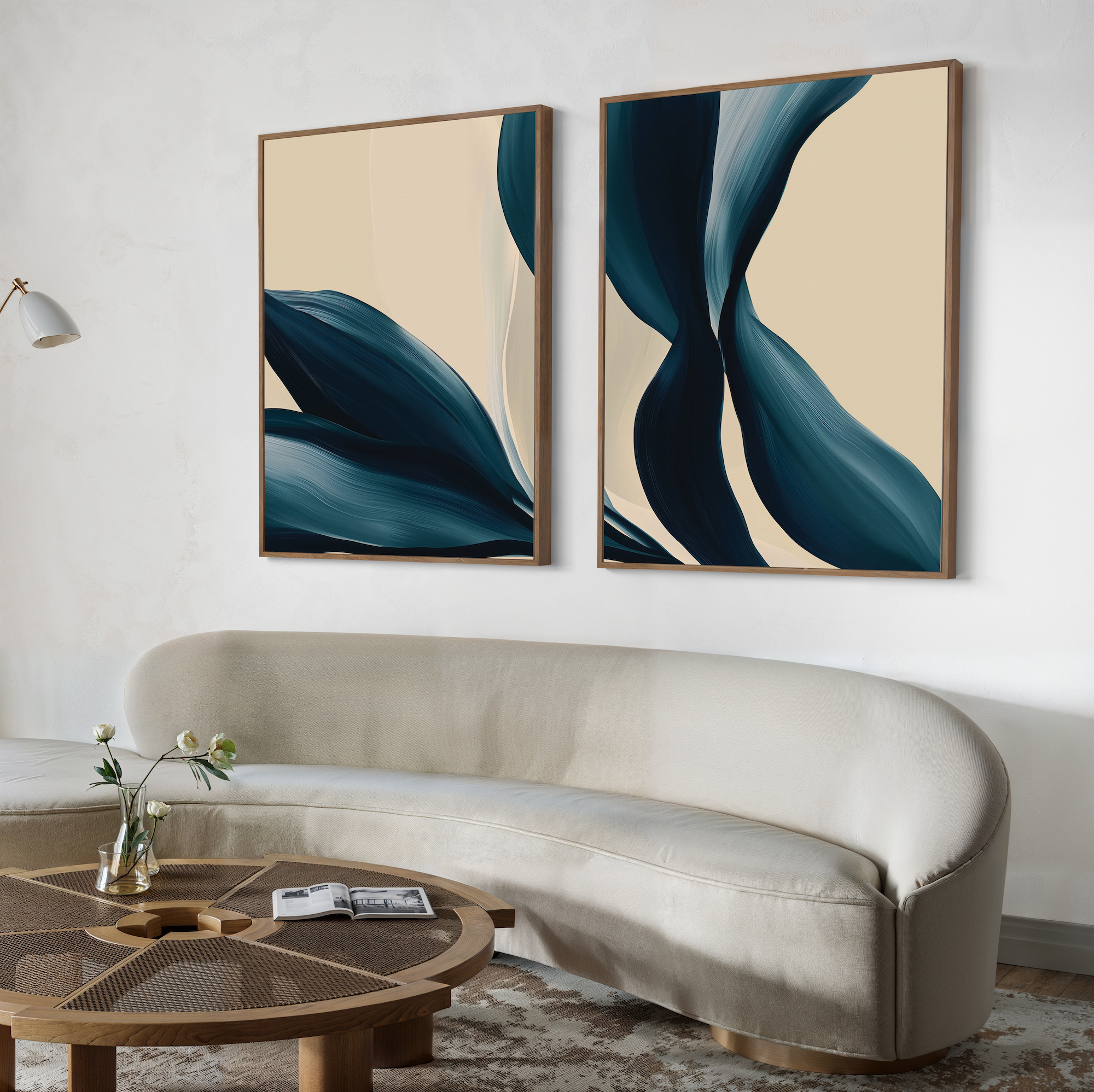 A modern living room features a curved beige sofa, a round wooden coffee table, and the Midnight Foliage Set of 2 abstract blue and cream paintings above the sofa. A white wall lamp and a flower vase complete the decor.