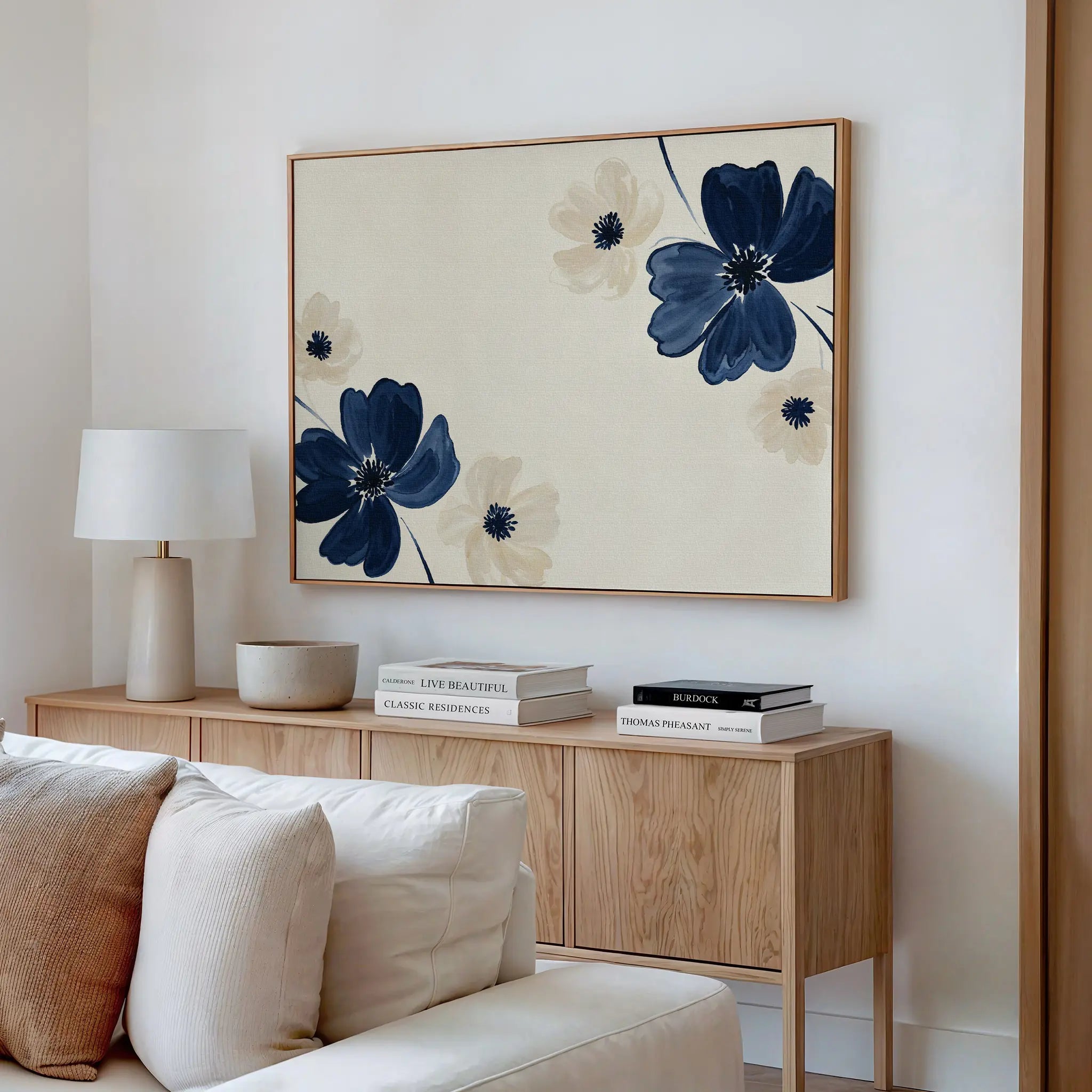 A cozy living room features a light wood sideboard, white table lamp, books, and neutral decor. Above the white couch with soft cushions hangs the Midnight Garden Canvas Art in blue and beige, adding a fresh touch to the space.