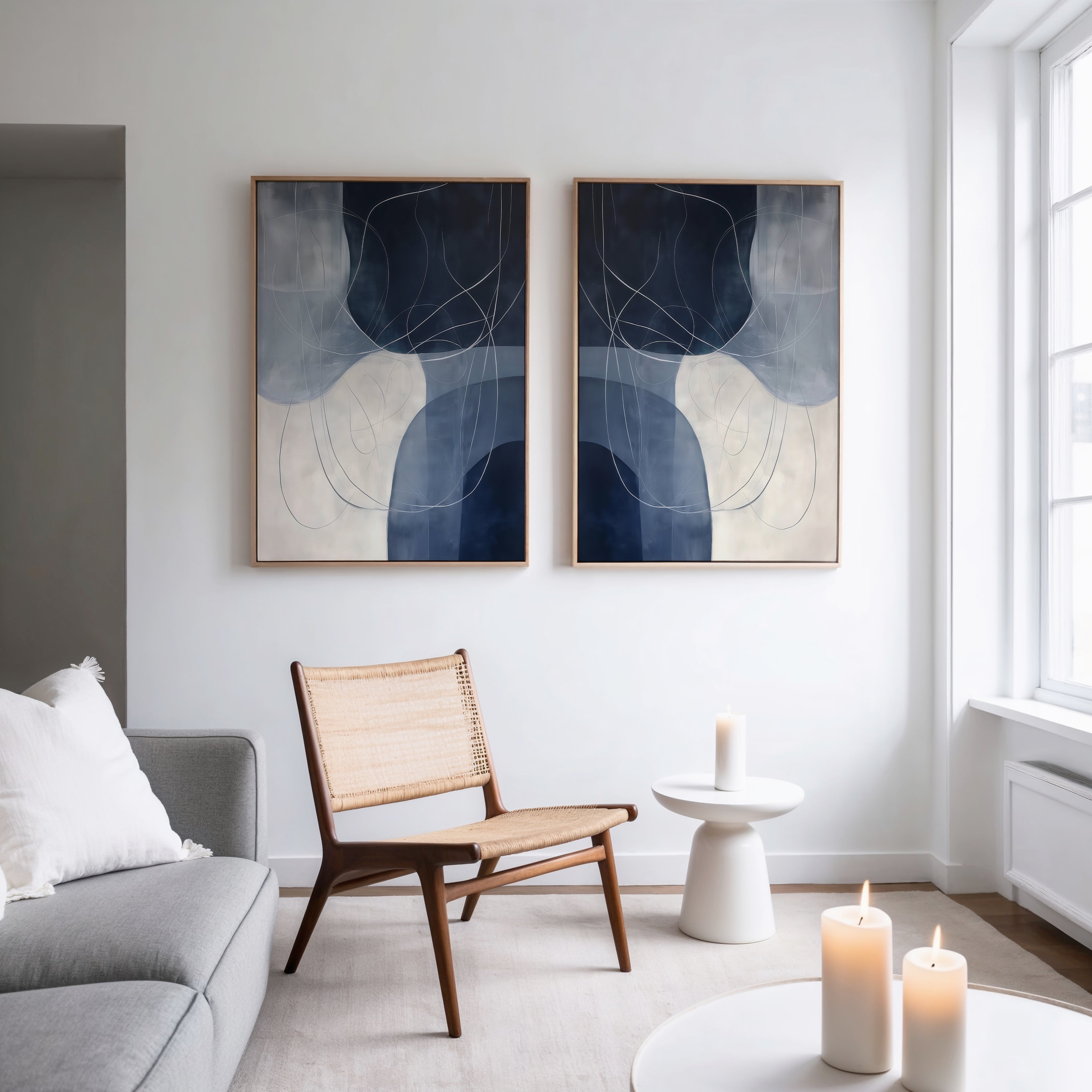 A modern living room with a light grey sofa, wooden chair with woven seat, small round white table, and candles. Above the chair hang the Midnight Lines Set of 2 minimalist blue and white art pieces, adding modern elegance to the space.
