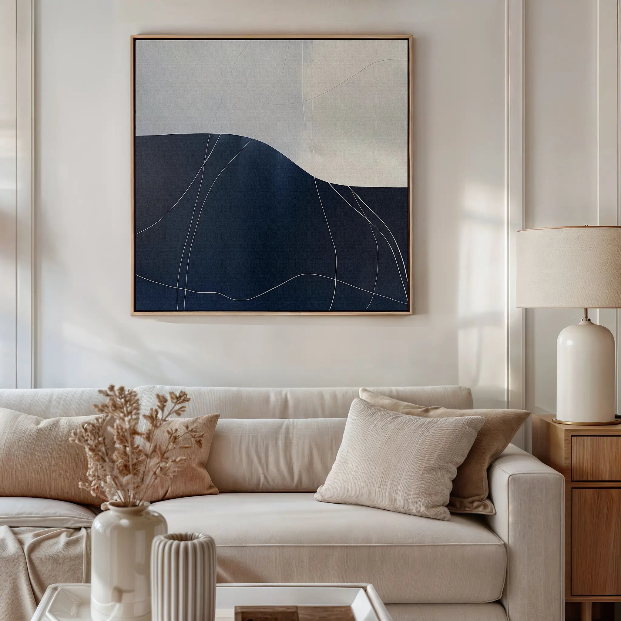 A modern living room featuring a beige sofa, neutral cushions, a wooden side table with a ceramic lamp, the Midnight Terrain Canvas Art in blue, white, and beige tones above the sofa, plus dried flowers and decor on the coffee table.