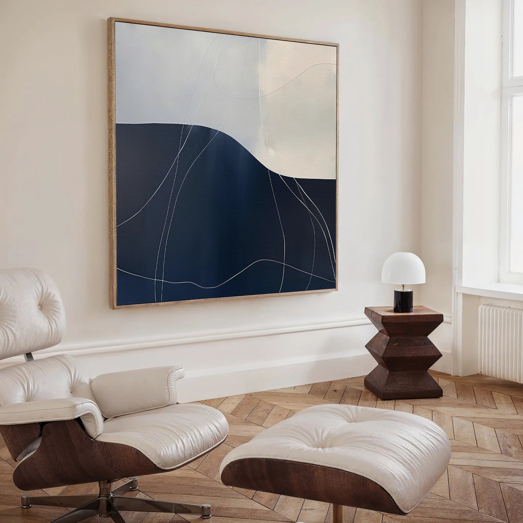 A modern living space featuring a white leather lounge chair with ottoman, a dark wood side table with a small white lamp, Midnight Terrain Canvas Art on the wall, and herringbone wooden floors for striking bedroom decor.