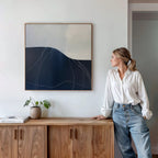 A woman in a white blouse and jeans leans against a wooden cabinet, looking at the Midnight Terrain Canvas Art above her. On the cabinet are a potted plant and books in a bright, modern room.