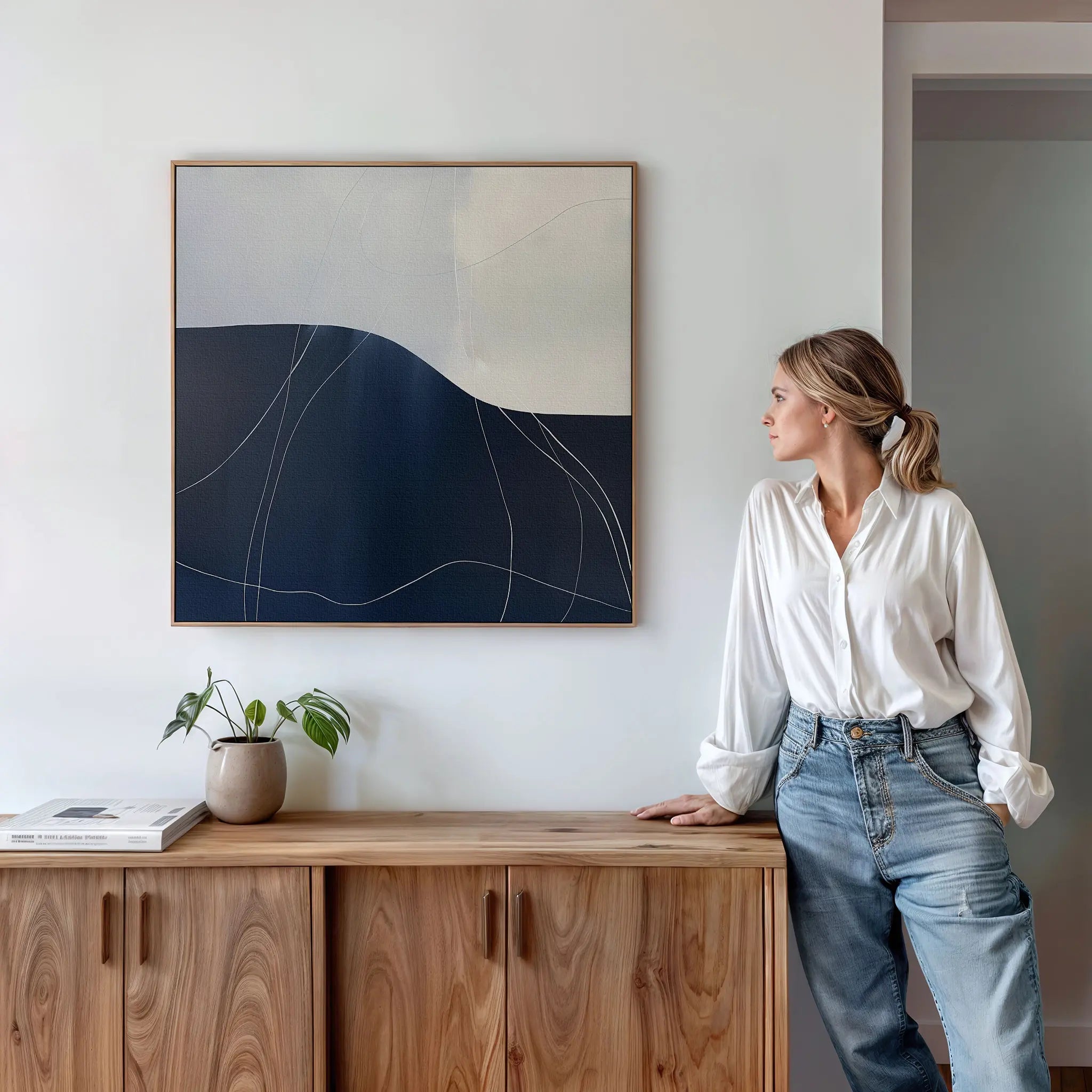 A woman in a white blouse and jeans leans against a wooden cabinet, looking at the Midnight Terrain Canvas Art above her. On the cabinet are a potted plant and books in a bright, modern room.
