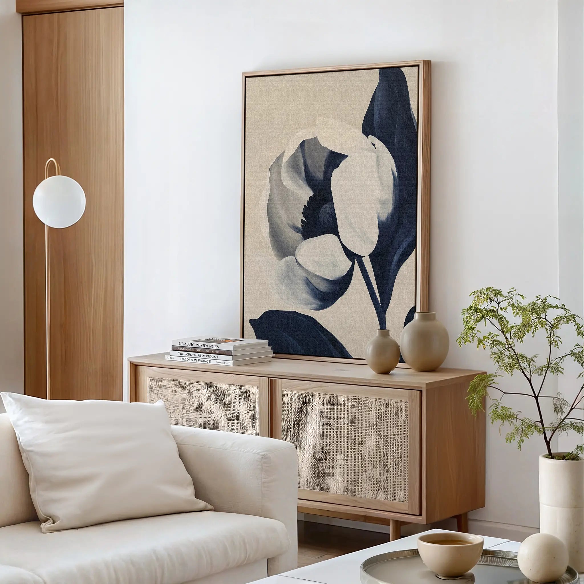A modern living room with neutral tones features a wooden sideboard, minimalist decor, a potted plant, and the Midnight Tulip Canvas Art in navy and beige above the cabinet. A white sofa with cushions completes the inviting space.