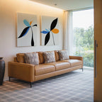 A modern living room with a tan leather sofa, three patterned cushions, the Minimal Flora Set of 2 wall art above, a large window with sheer curtains, and a decorative vase on the floor.
