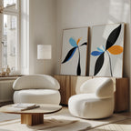 A modern living room with two rounded white chairs, a low wooden coffee table, and the Minimal Flora Set of 2—abstract floral art pieces in black, blue, and orange—displayed on a wooden sideboard. Sunlight pours through large windows.