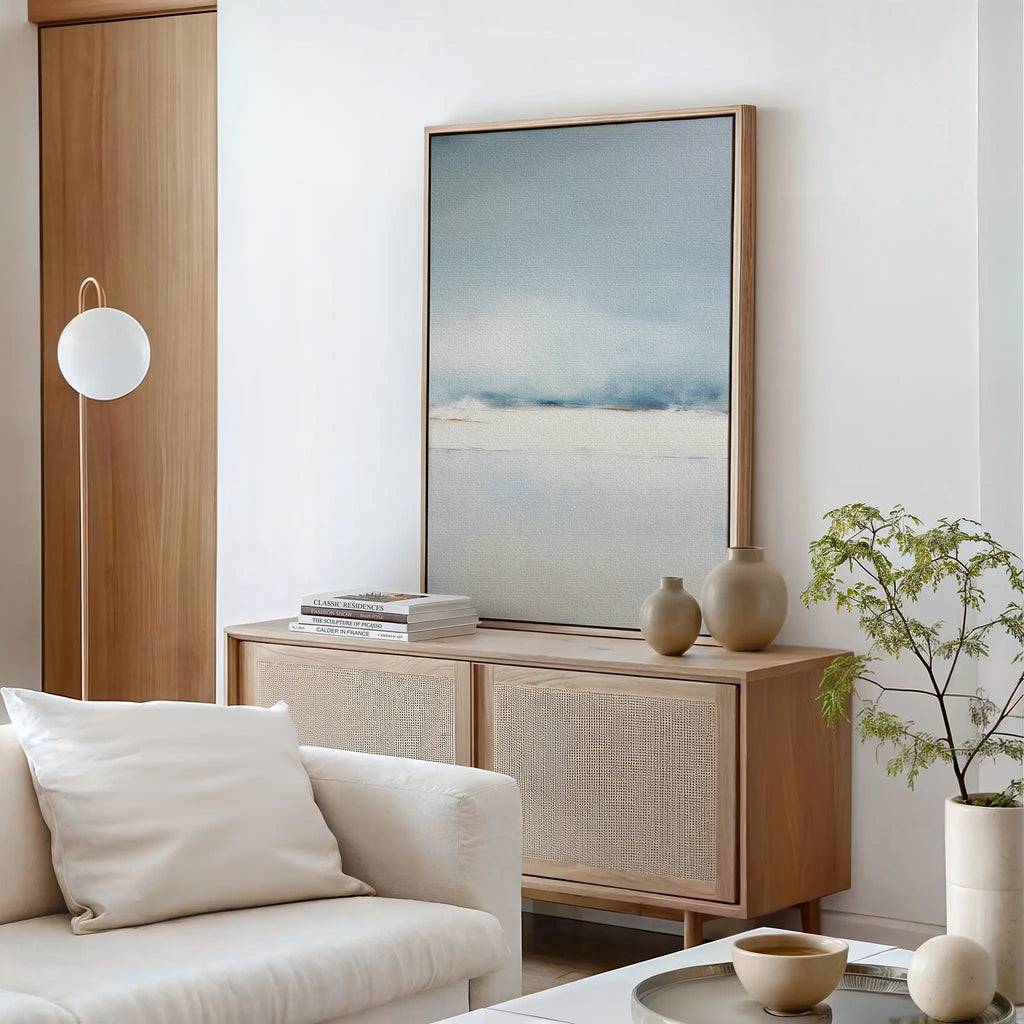 A modern living room with a cream sofa, light wood sideboard, minimalist vases, a potted plant, books, a round wall lamp, and Misty Morning Canvas Art – Vertical creates a tranquil atmosphere in soft blue and beige tones.