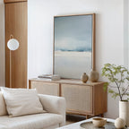 A modern living room with a cream sofa, light wood sideboard, minimalist vases, a potted plant, books, a round wall lamp, and Misty Morning Canvas Art – Vertical creates a tranquil atmosphere in soft blue and beige tones.