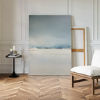 The Misty Morning Canvas Art – Vertical, featuring soft blue and white tones, leans against a paneled wall to create tranquility. Nearby, a black round side table with candles and books sits beside a simple wooden chair with a white cushion.