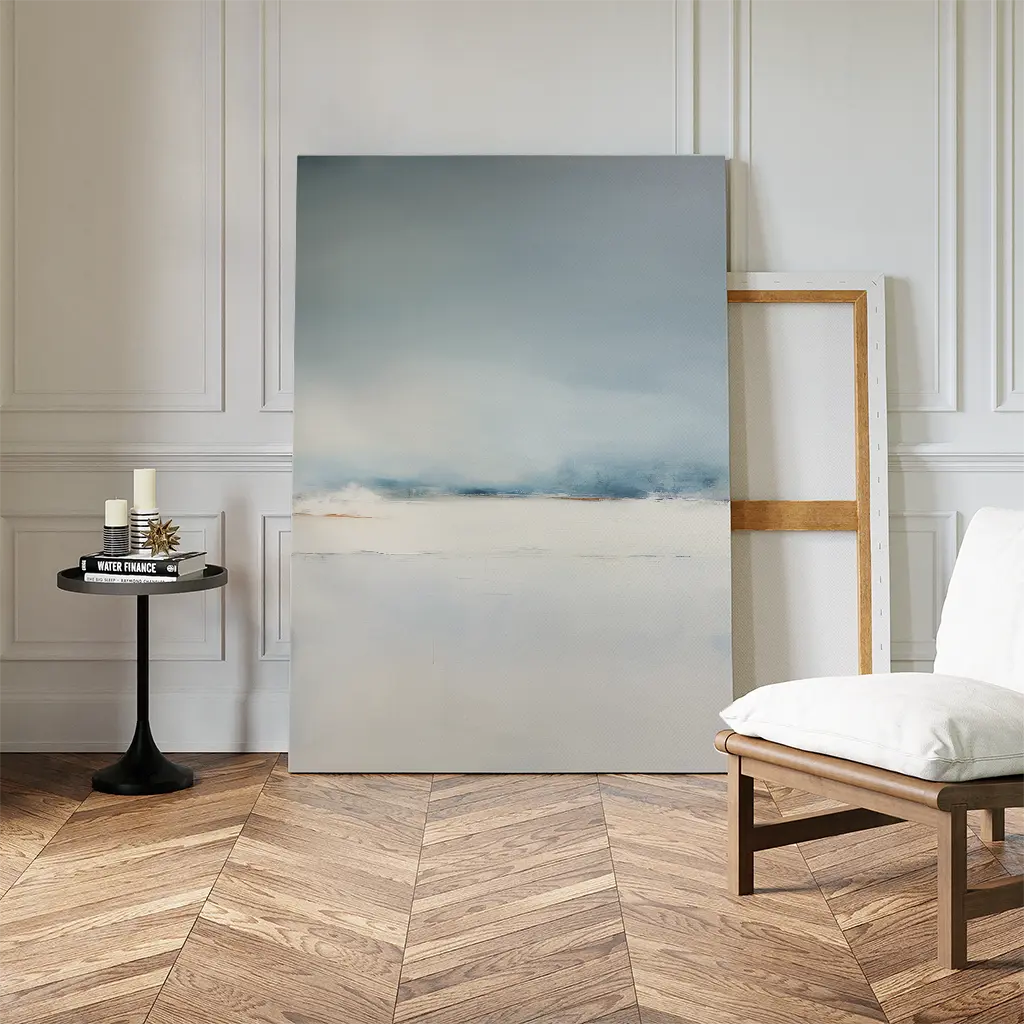 The Misty Morning Canvas Art – Vertical, featuring soft blue and white tones, leans against a paneled wall to create tranquility. Nearby, a black round side table with candles and books sits beside a simple wooden chair with a white cushion.