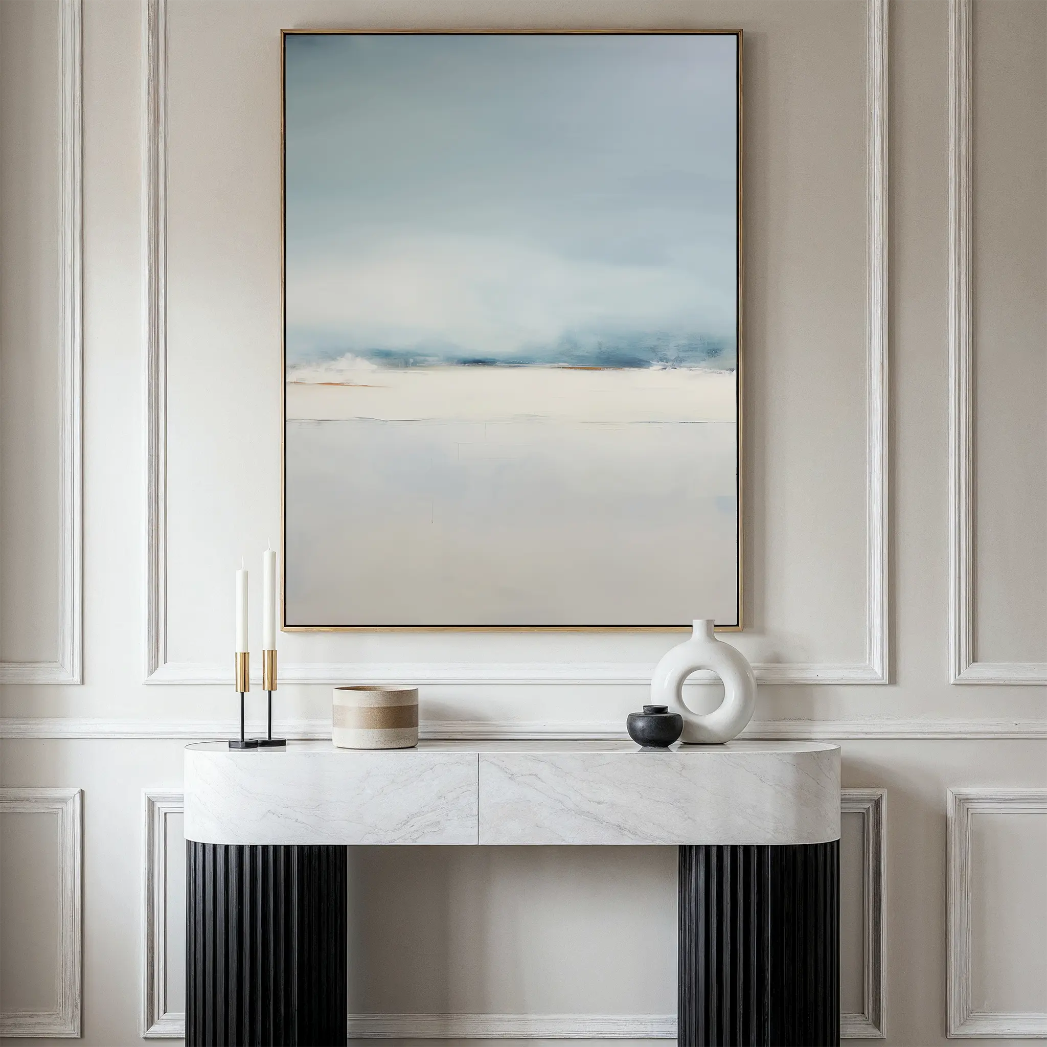 A modern console table with decor stands against a cream paneled wall; above it hangs the Misty Morning Canvas Art – Vertical, showcasing a serene landscape in soft blue, gray, and beige tones for a tranquil atmosphere.