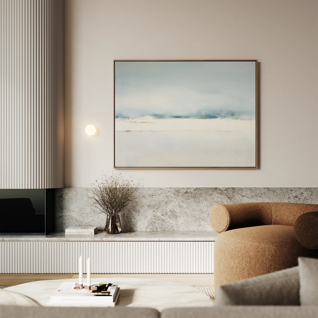 A modern living room featuring the Misty Morning Canvas Art – Horizontal in soft blue and white, a beige curved sofa, marble accents, a small vase with dried flowers, and a stack of books with candles on a low table.