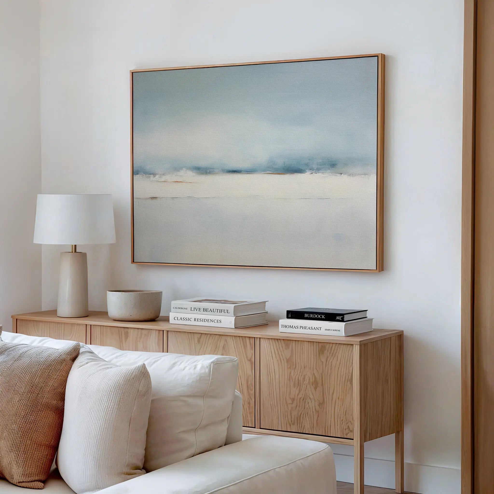 A minimalist living room features a light wooden sideboard with books, a lamp, and a bowl. Above, Misty Morning Canvas Art – Horizontal hangs on the white wall. A cream sofa with cushions is in the foreground.
