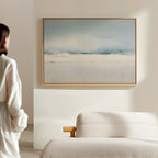 A person stands in a minimalist room with light walls, admiring the Misty Morning Canvas Art – Horizontal in blue, white, and beige above a cream sofa, bringing a touch of coastal-inspired decor.