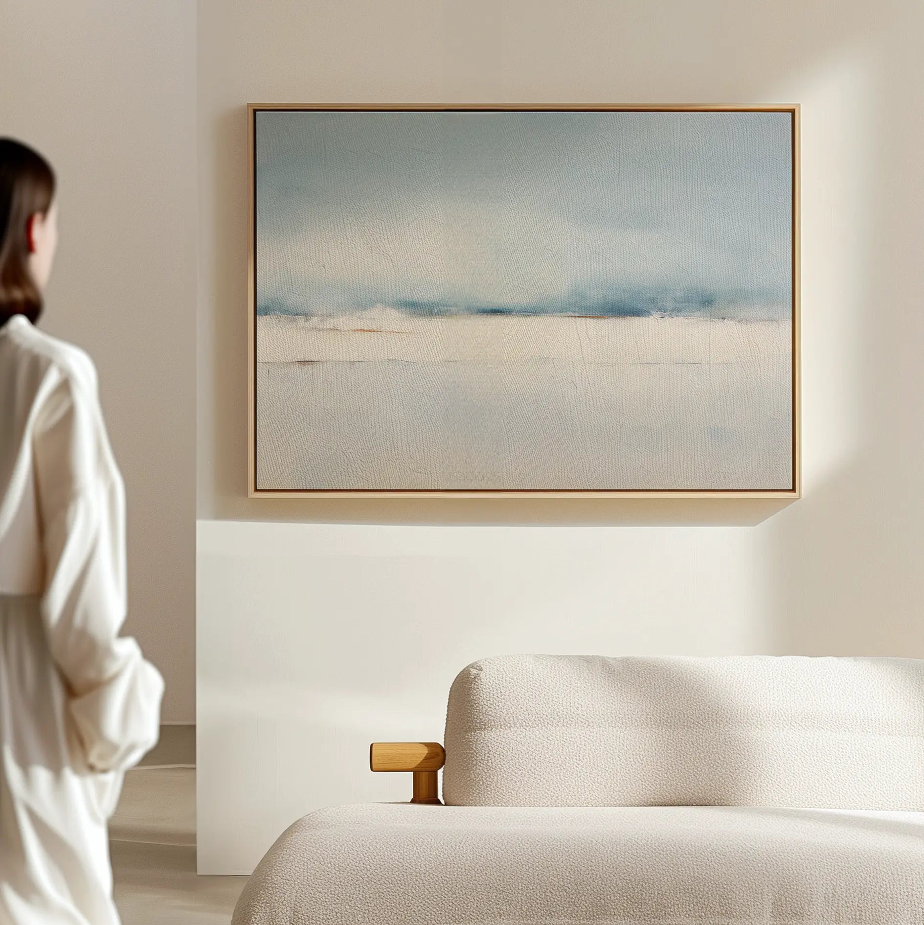 A person stands in a minimalist room with light walls, admiring the Misty Morning Canvas Art – Horizontal in blue, white, and beige above a cream sofa, bringing a touch of coastal-inspired decor.