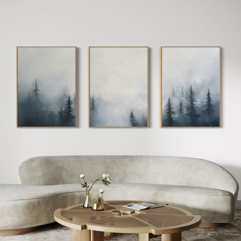 A modern living room with a curved beige sofa, round wooden coffee table with books and a vase, and the Misty Pines Set of 3 wall art—framed paintings of misty pine trees in Nordic Park style—hanging on a white wall.