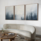 A minimalist living room features a curved cream sofa, round wooden coffee table, and the Misty Pines Set of 3 wall art above the sofa. A small vase with flowers on the table enhances the serene Nordic Park ambience.