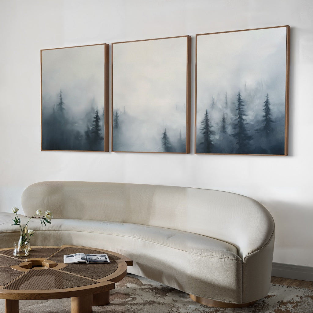 A minimalist living room features a curved cream sofa, round wooden coffee table, and the Misty Pines Set of 3 wall art above the sofa. A small vase with flowers on the table enhances the serene Nordic Park ambience.
