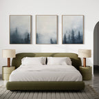 A modern bedroom features an olive green bed, white bedding, beige lamps on round nightstands, and the Misty Pines Set of 3—three framed paintings of misty pine trees—displayed above the bed.
