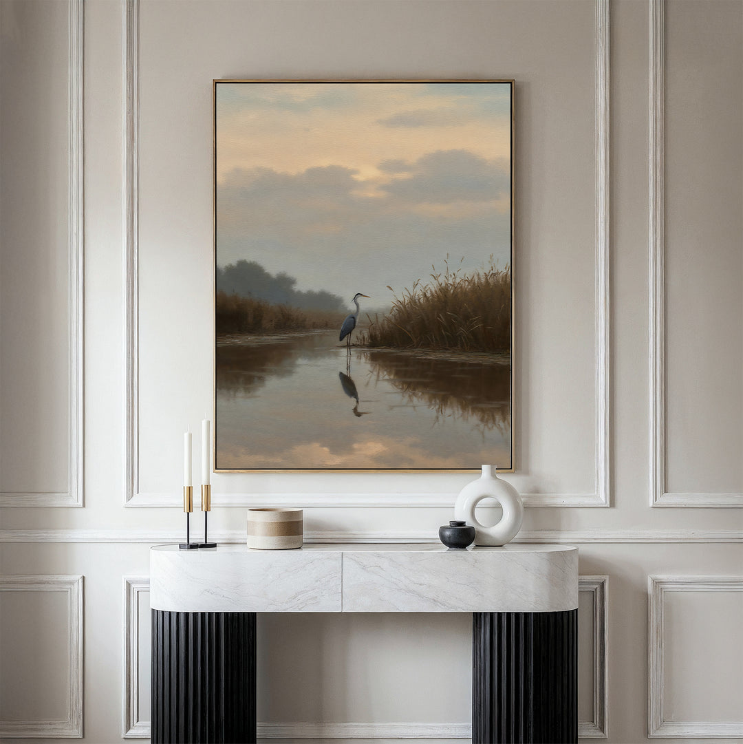A modern console table with decorative objects stands against a white paneled wall. Above it hangs the Misty Reflection Canvas Art, featuring a serene heron by a river at sunset, with reeds and its reflection in the water.