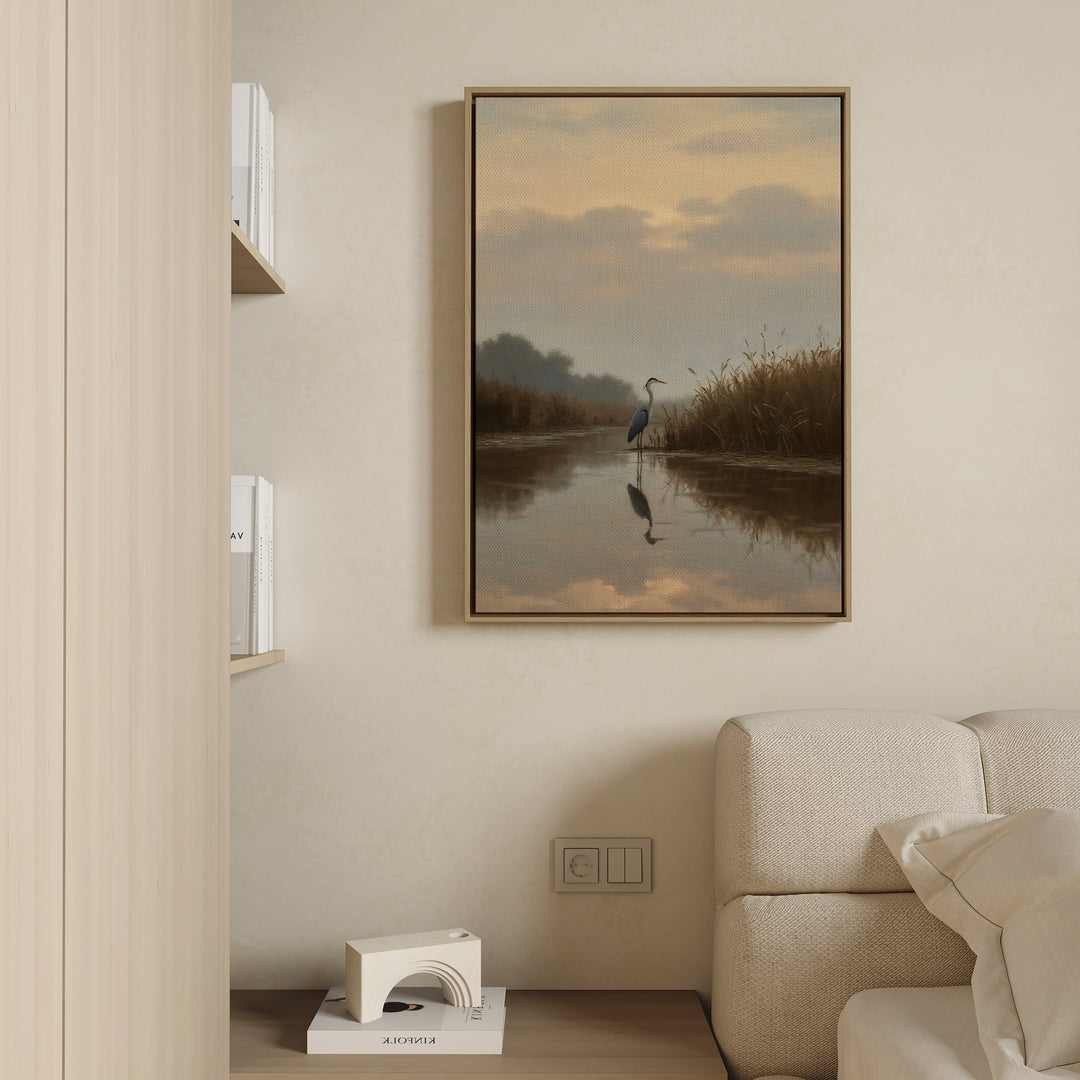 The Misty Reflection Canvas Art, featuring a tranquil heron by a calm river, hangs above a light sofa on a beige wall. Built-in shelves nearby display books and a geometric decor piece sits on the lower shelf.