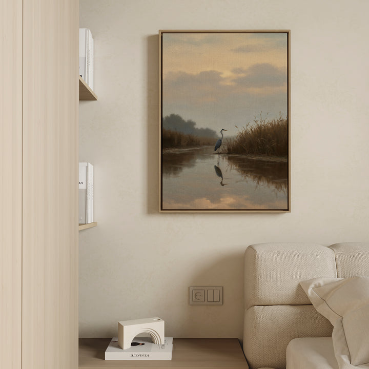 The Misty Reflection Canvas Art, featuring a tranquil heron by a calm river, hangs above a light sofa on a beige wall. Built-in shelves nearby display books and a geometric decor piece sits on the lower shelf.