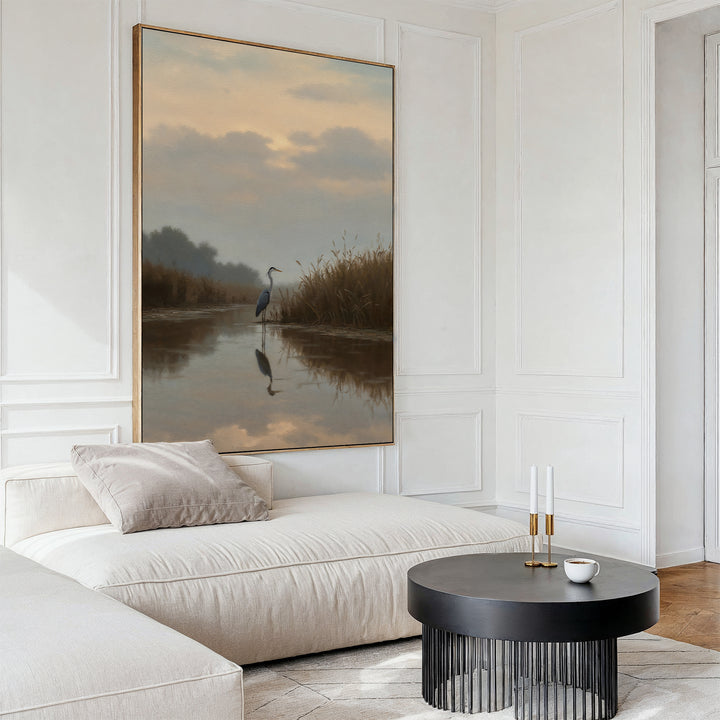 A modern living room featuring a white sectional sofa, round black coffee table, and Misty Reflection Canvas Art depicting a serene heron in a calm marsh. Neutral tones and minimalist decor enhance the tranquil atmosphere.