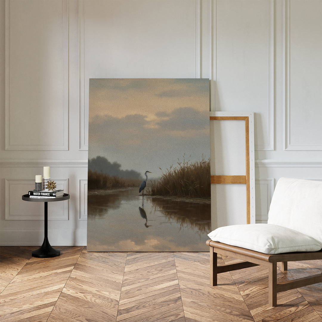 The Misty Reflection Canvas Art, featuring a heron by a sunset river, leans against a white paneled wall in a minimalist room with wood floors, a cushioned chair, and a small round table holding candles and books.