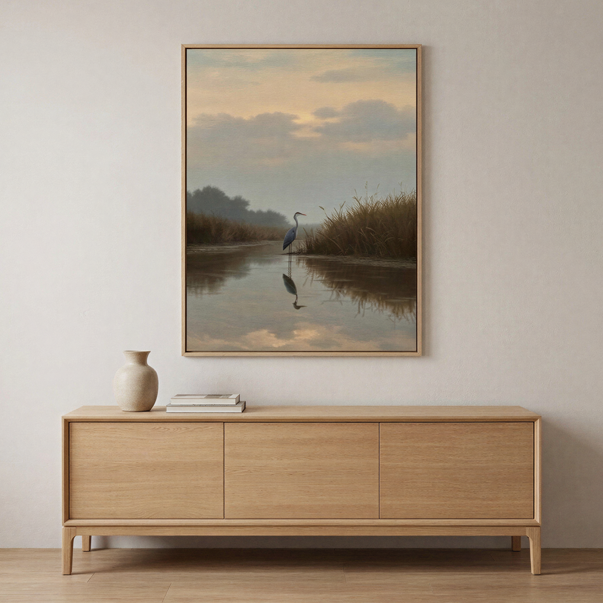 A minimalist room with a light wood sideboard, ceramic vase, and books. Above, Misty Reflection Canvas Art—a framed heron by calm water at sunset—complements the serene, neutral aesthetic.