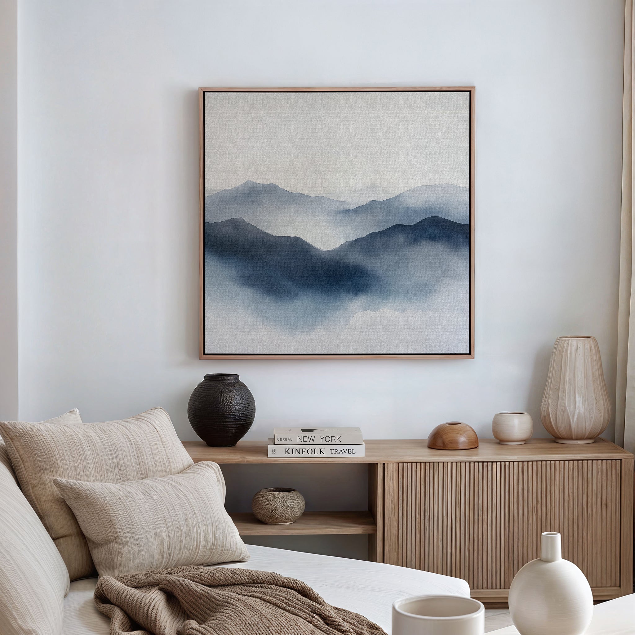 A serene living room with neutral tones, a beige sofa, a wooden sideboard decorated with vases and books, and the Misty Ridge Canvas Art—an abstract depiction of blue mountains—featured prominently on the wall.