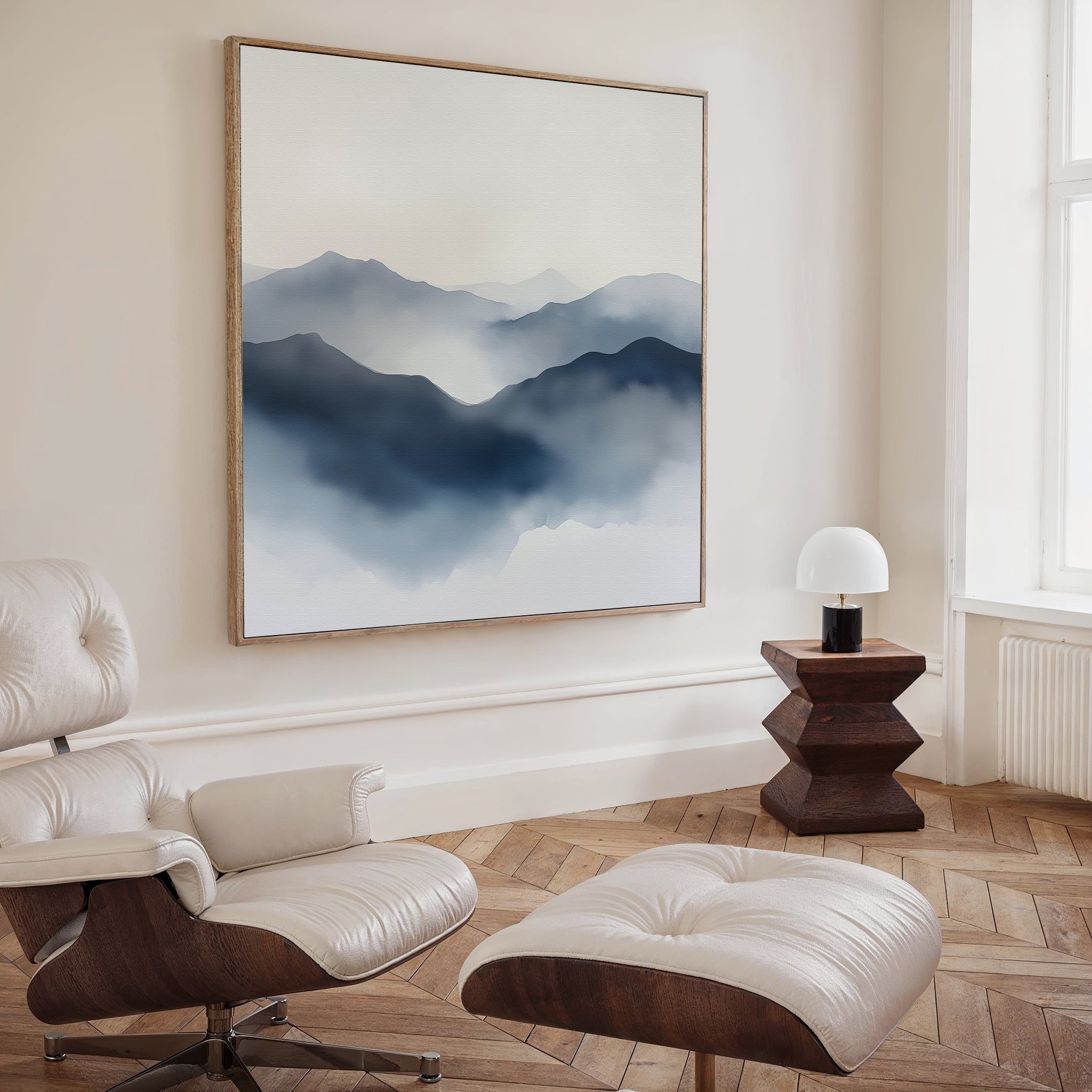 A modern, minimalist living room features light wood herringbone floors, a white leather lounge chair and ottoman, a geometric wooden side table with a small white lamp, and Misty Ridge Canvas Art as the focal point on the wall.