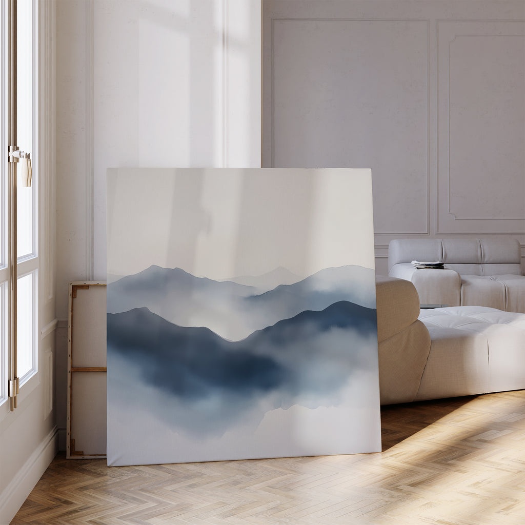 The Misty Ridge Canvas Art, a large minimalist landscape featuring misty blue mountains, leans against the wall in a bright modern living room with a white couch, parquet flooring, and sunlight streaming through tall windows.