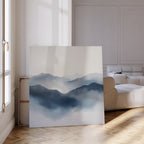 The Misty Ridge Canvas Art, a large minimalist landscape featuring misty blue mountains, leans against the wall in a bright modern living room with a white couch, parquet flooring, and sunlight streaming through tall windows.