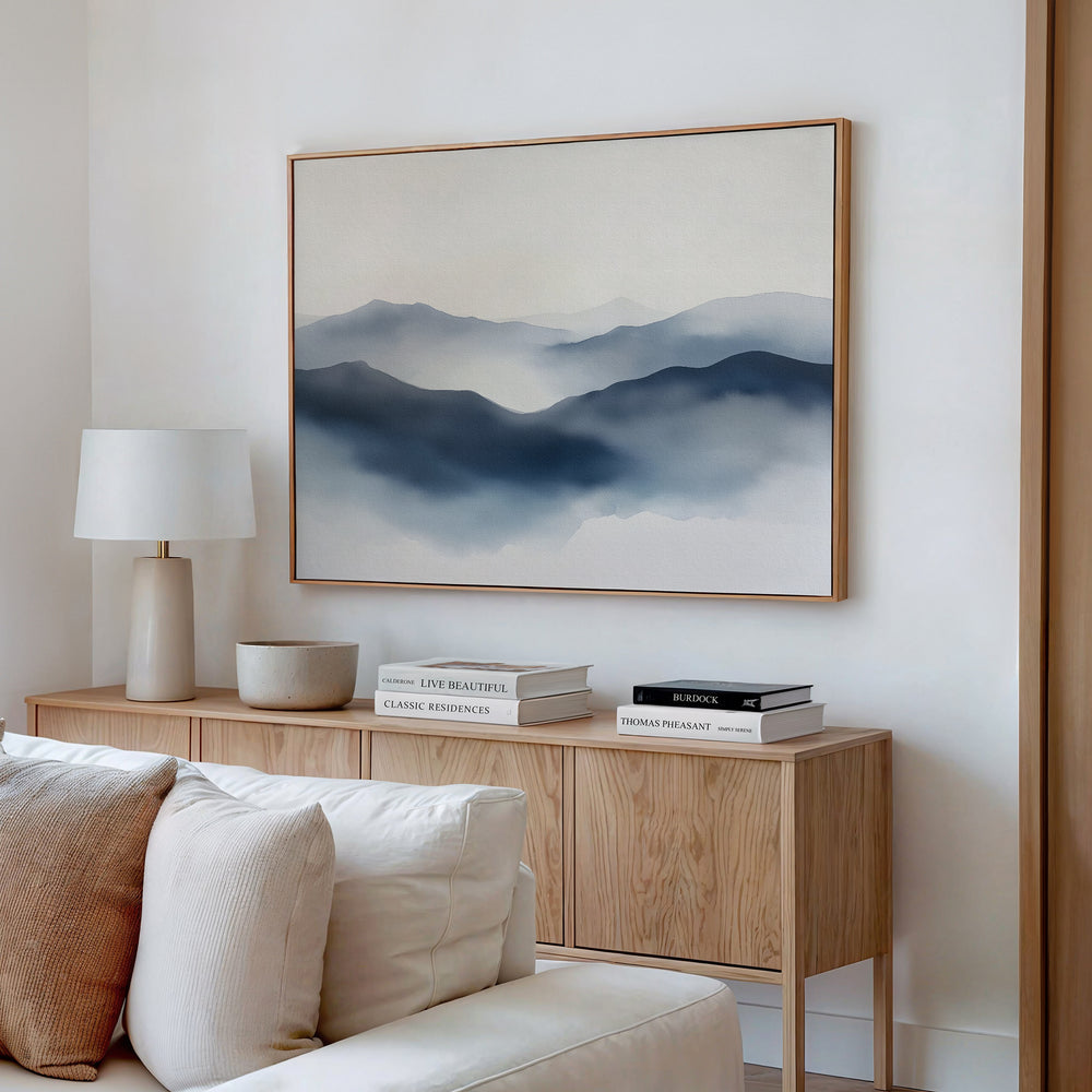 A minimalist living room featuring a light wood sideboard, stacked books, a white table lamp, a bowl, beige sofa, and Misty Ridge Canvas Art – Horizontal depicting blue-gray misty mountains on a white wall.