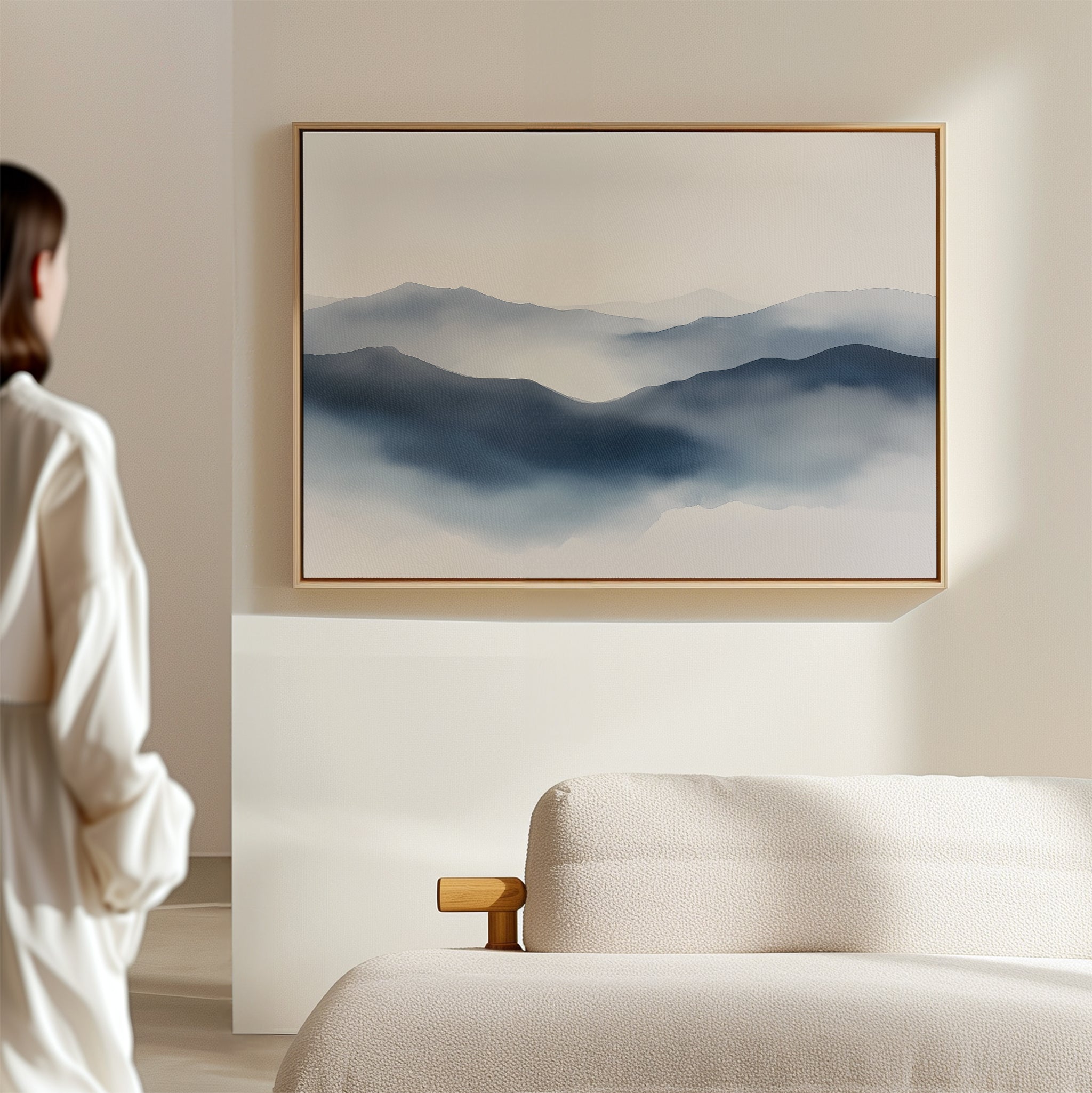 A woman stands in a minimal, light-filled room with a soft beige couch and the Misty Ridge Canvas Art – Horizontal depicting blue mountains hanging on the wall.