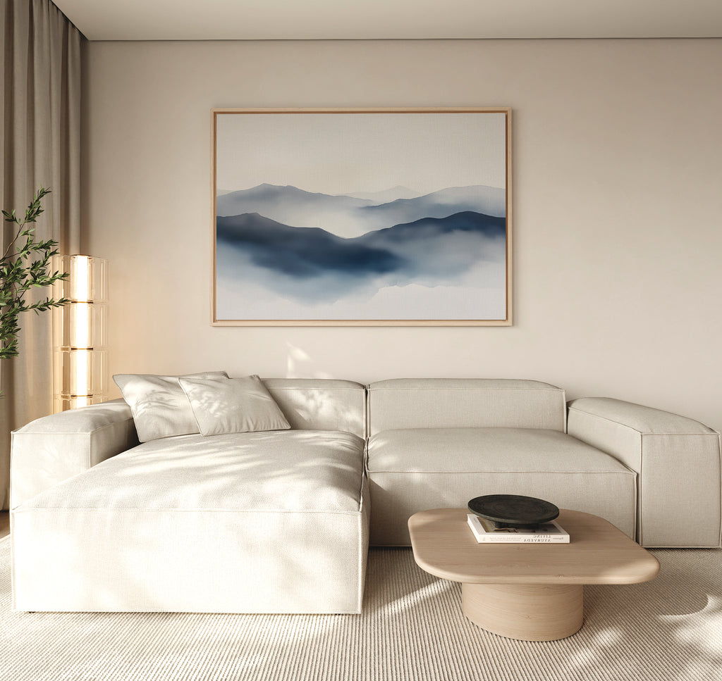 A minimalist living room with a light beige sectional sofa, low wooden coffee table, potted plant, and a Misty Ridge Canvas Art – Horizontal depicting blue mountains on a neutral wall.