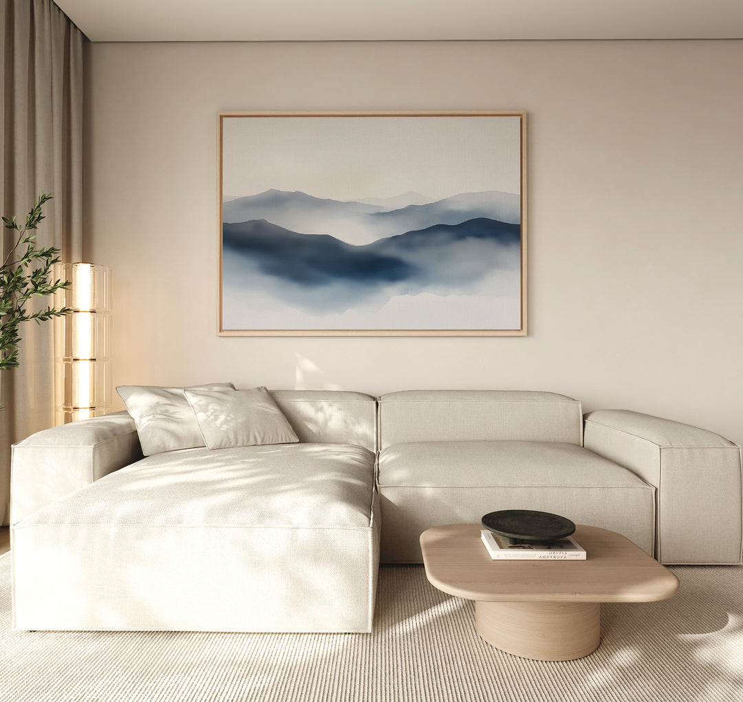 A minimalist living room with a light beige sectional sofa, low wooden coffee table, potted plant, and a Misty Ridge Canvas Art – Horizontal depicting blue mountains on a neutral wall.