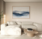 A minimalist living room with a light beige sectional sofa, low wooden coffee table, potted plant, and a Misty Ridge Canvas Art – Horizontal depicting blue mountains on a neutral wall.