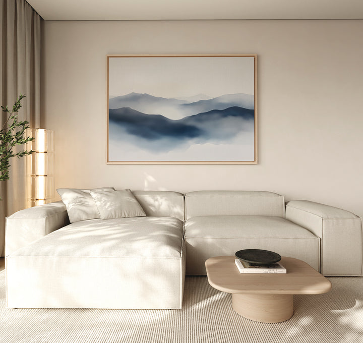 A minimalist living room with a light beige sectional sofa, low wooden coffee table, potted plant, and a Misty Ridge Canvas Art – Horizontal depicting blue mountains on a neutral wall.