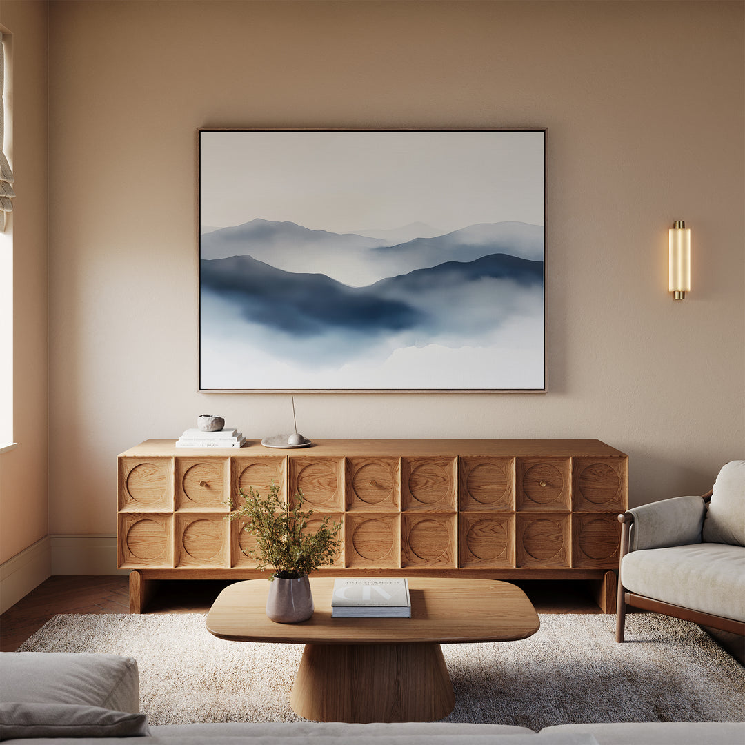 A cozy living room featuring a wooden sideboard with circular patterns, a modern light fixture, a beige sofa, a low wooden coffee table, and the Misty Ridge Canvas Art – Horizontal depicting mountains on the wall.